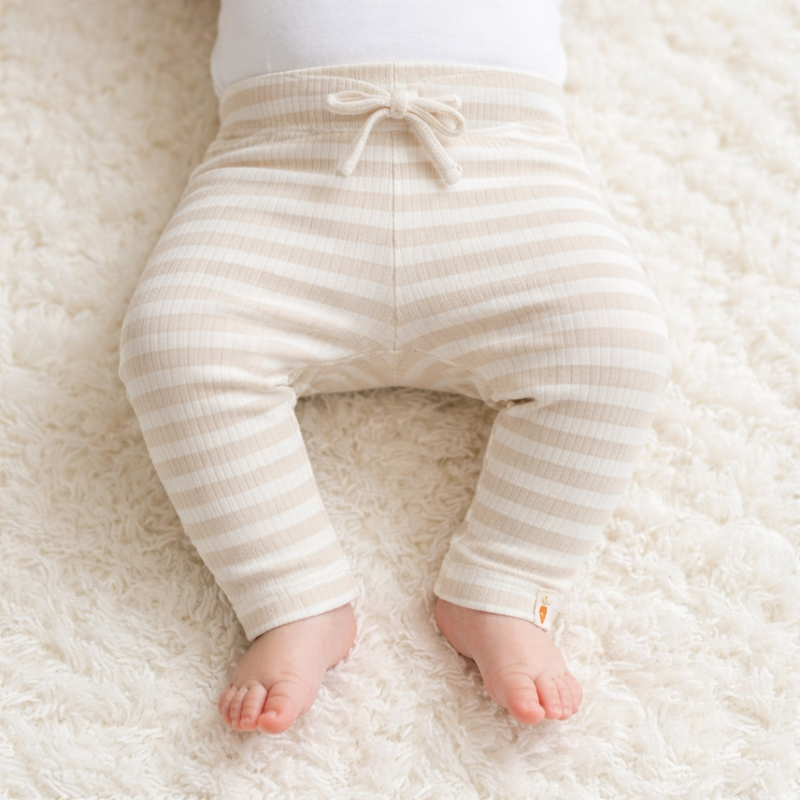 Baby trägt eine beige gestreifte Ripp Leggings mit Bindeband und liegt auf einer weichen Decke
