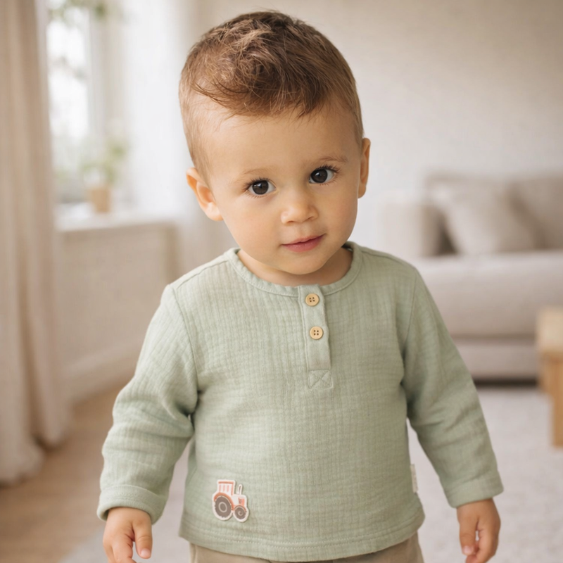 Kleiner Junge trägt ein langärmeliges grünes Musselin Shirt mit Knopfleiste und Traktor Motiv und steht in einem hellen Wohnraum