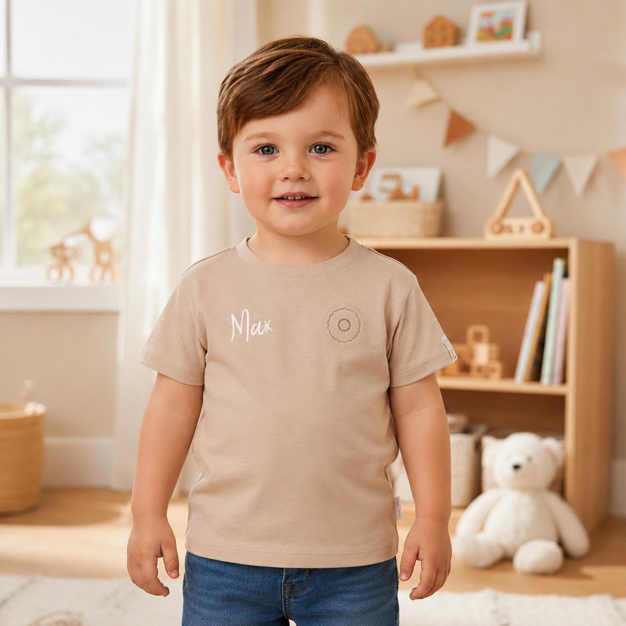 Kleinkind trägt ein personalisiertes beiges Kinder-T-Shirt mit dem Namen Max auf der Brust und steht in einem hellen Kinderzimmer.