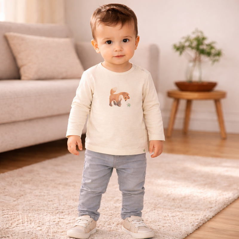 Kleiner Junge trägt ein beiges langärmeliges Shirt mit Hund Motiv sowie eine graue Hose und steht auf einem weichen Teppich in einem hellen Wohnzimmer