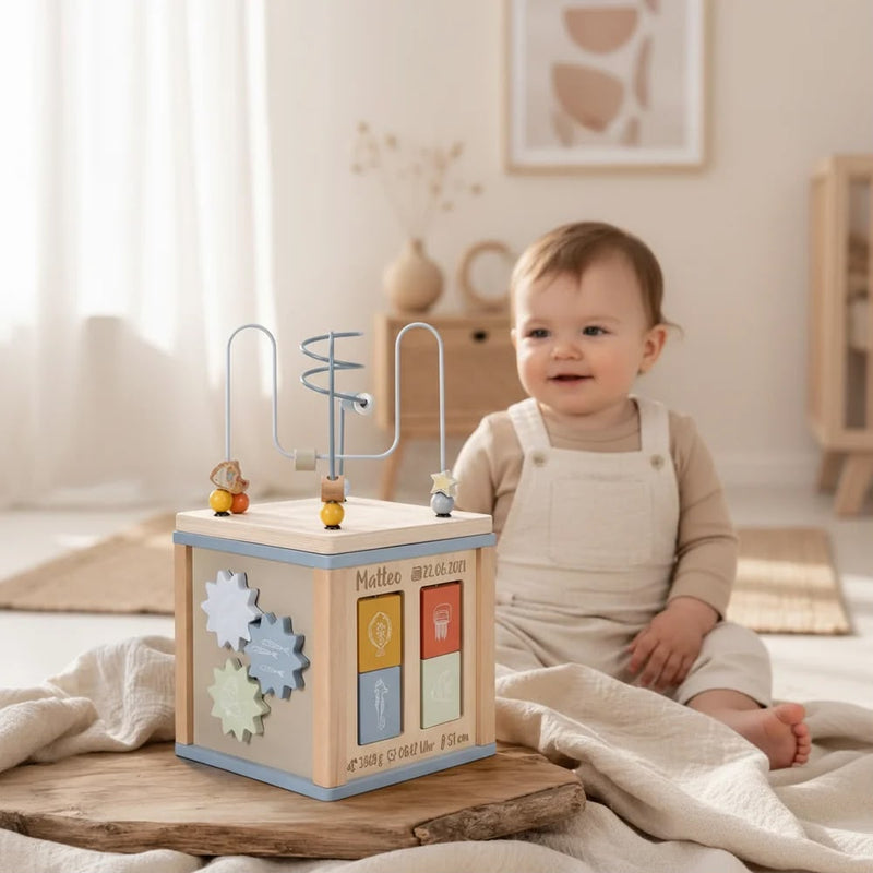 Personalisierter Motorikwürfel aus Holz mit Name, Geburtsdatum, Geburtszeit, Gewicht und Größe als individuelles Geschenk zur Geburt. Hochwertiges Montessori-Holzspielzeug für Babys ab 12 Monaten mit Motorikschleife und Zahnrädern in einer natürlichen Babyzimmer-Umgebung.
