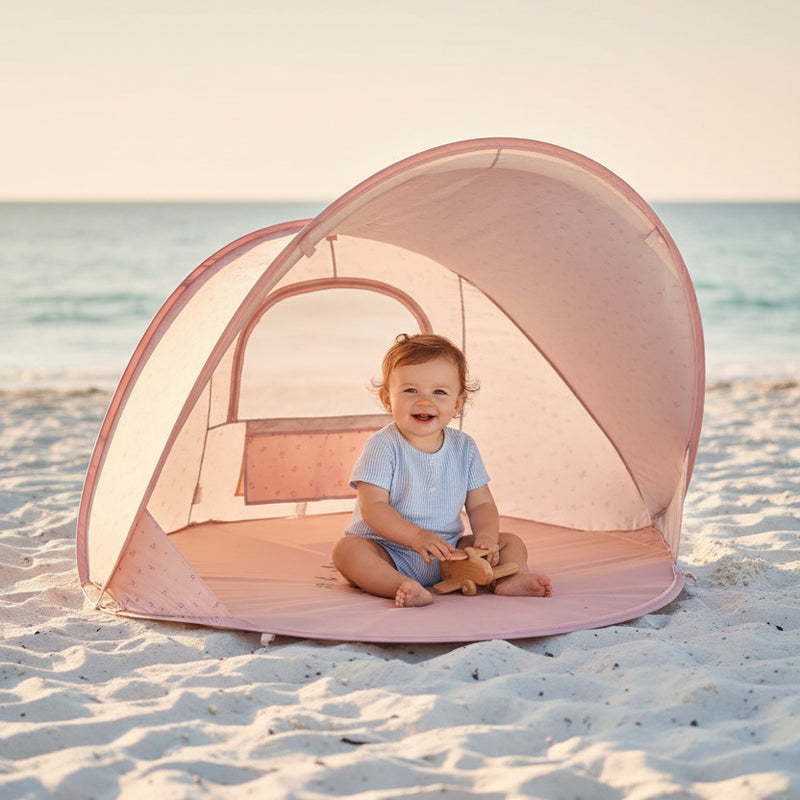 Baby sitzt im Schatten in einer pinken Strandmuschel am Strand – UV Schutz Pop Up Strandzelt für Babys und Kinder im Sommerurlaub